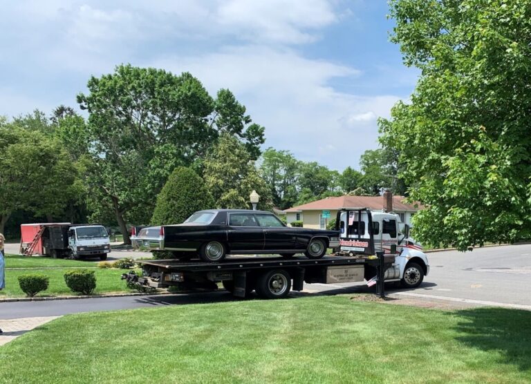 A blue 4 door Cadillac on a flatbed truck driving down a driveway on its way to a DIY classic car restoration